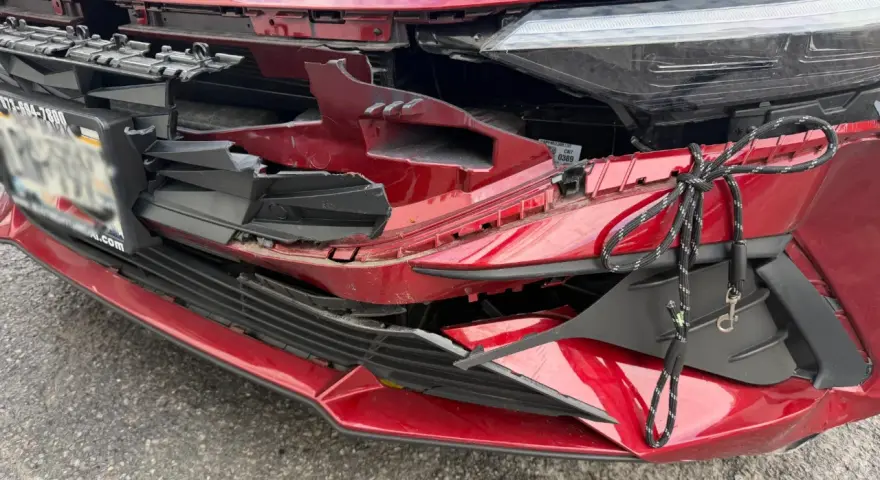 A red car's front bumper is severely damaged, with broken plastic and a rope tied to it. This damage might be from hail, and the owner is likely wondering how do you repair hail damage on a car.