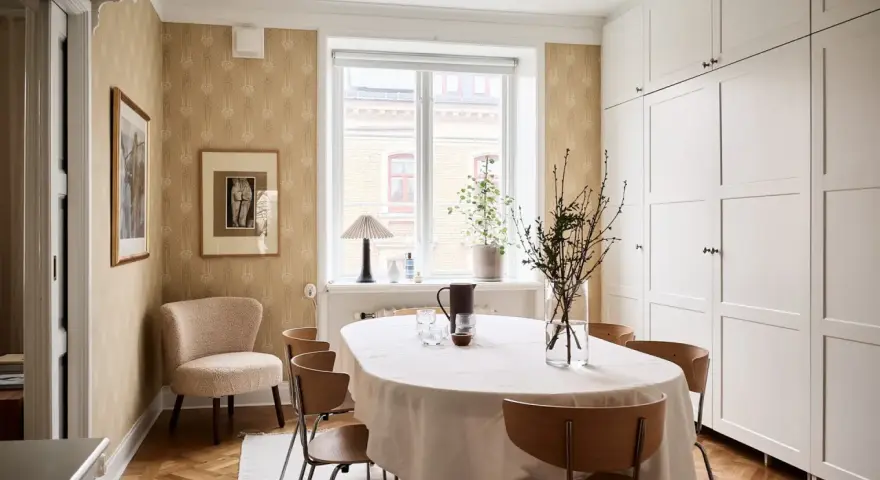 Comedor con mesa cubierta, sillas de madera y pared con papel tapiz dorado. Armarios blancos y suelo de parqué.