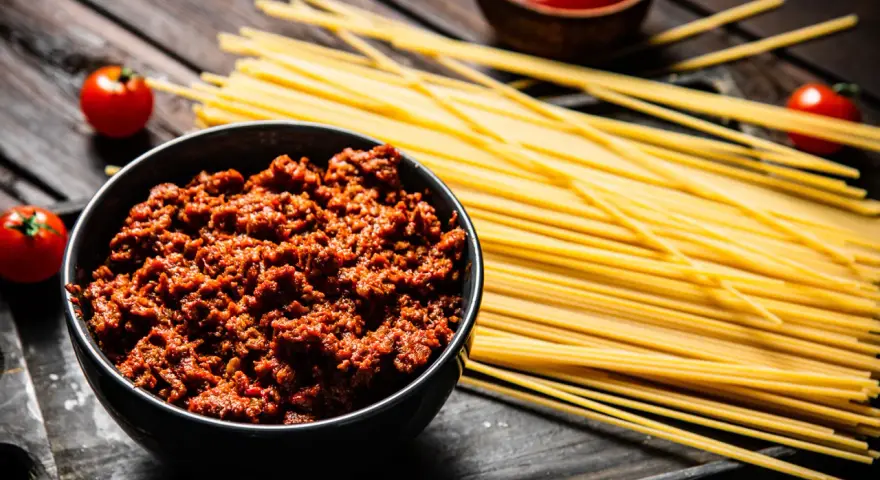 Un tazón de carne para espaguetis a la boloñesa, junto a pasta cruda, tomates y salsa.