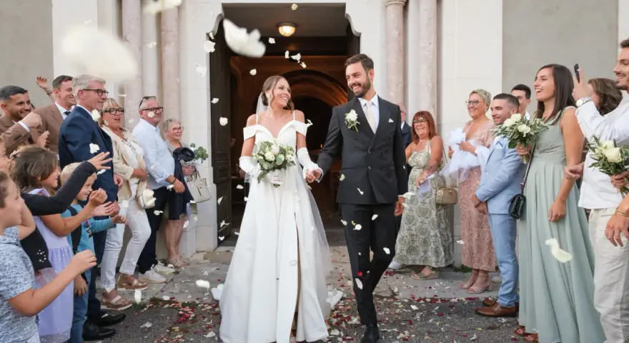 Photographie de mariage : le couple sort de l'église sous une pluie de pétales, entouré de leurs invités joyeux.