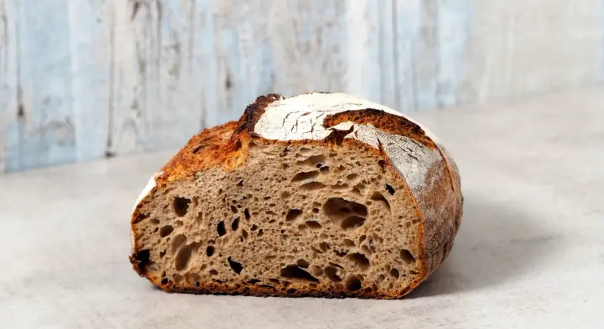 Ein halbes, knuspriges schnelles Sauerteigbrot mit großer Krume und rustikaler Kruste liegt auf einem hellen Untergrund vor einer hellblauen Holzvertäfelung.