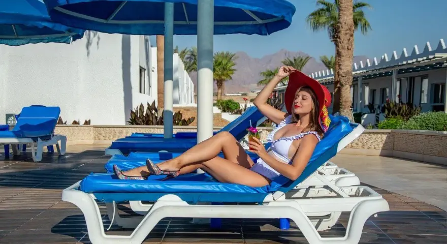 Kobieta w białym bikini i czerwonym kapeluszu relaksuje się na leżaku z drinkiem, ciesząc się wakacjami w stylu ultra all inclusive.