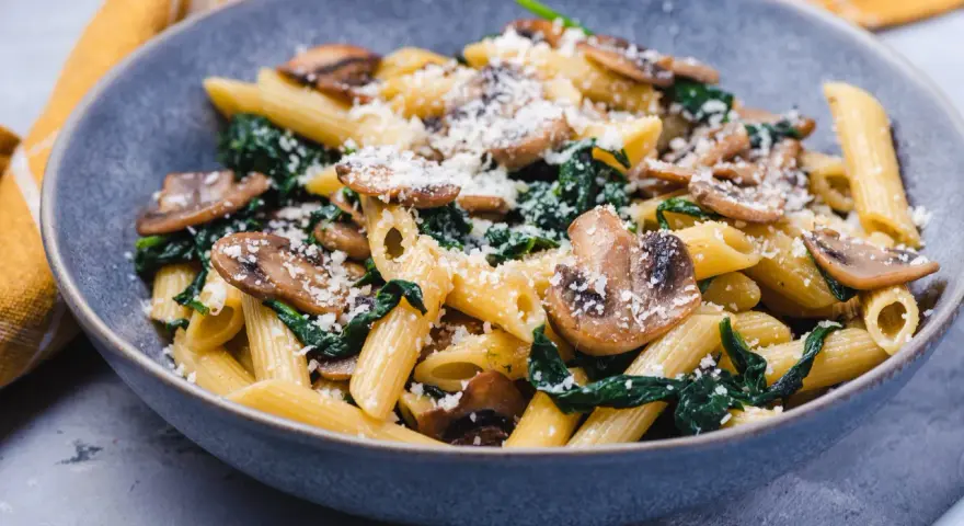 Penne z pieczarkami i szpinakiem, posypane parmezanem, w szarej misce.