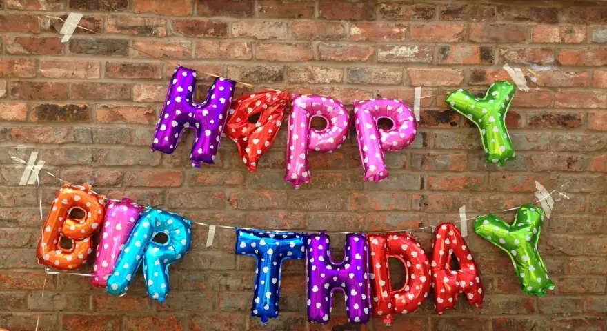 Luftballons in Buchstabenform bilden "HAPPY BIRTHDAY" an einer Ziegelwand.