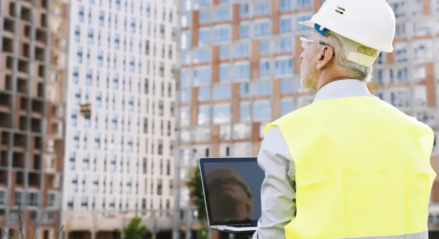 Ingeniero con casco y chaleco reflectante revisa un portátil frente a edificios en construcción. A pesar de un posible **ite desfavorable**, su mirada es decidida.