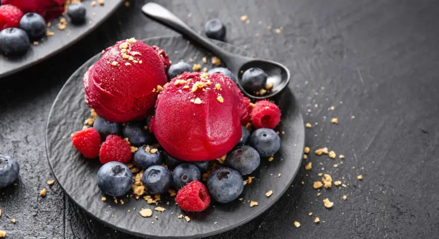 Zwei Kugeln Campari Sorbet mit frischen Beeren und Streuseln auf einem schwarzen Teller.