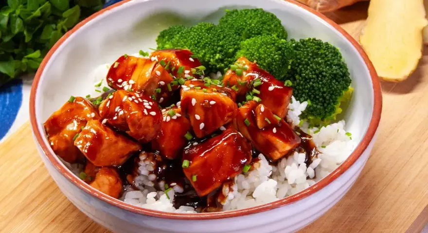 Miseczka ryżu z kawałkami lśniącego lososia teriyaki, posypanego sezamem i szczypiorkiem, obok parujących różyczek brokułów.