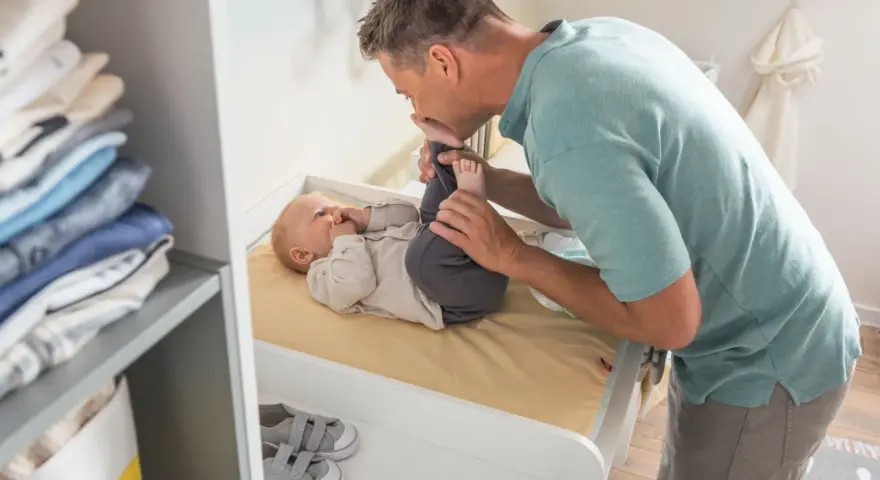 Ojciec całuje stópki dziecka na przewijaku. Wymiary przewijaka są idealne do pielęgnacji malucha. Obok stosik ubranek.