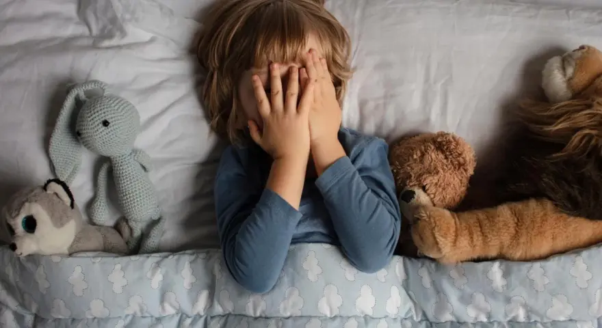 Kind versteckt Gesicht, umgeben von Kuscheltieren. Die Angst um das Kind ist spürbar, es sucht Trost bei seinen Freunden.