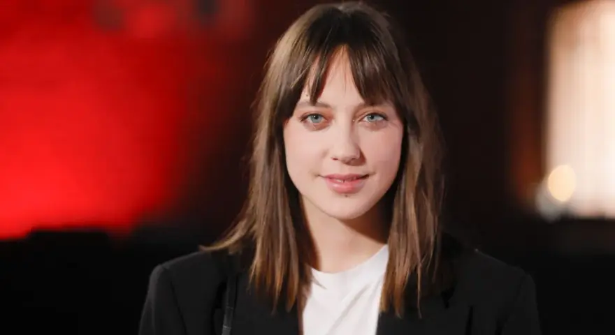 Maria Sobocińska z niebieskimi oczami i grzywką, ubrana w czarną marynarkę i białą koszulkę, uśmiecha się lekko na tle czerwonego światła.
