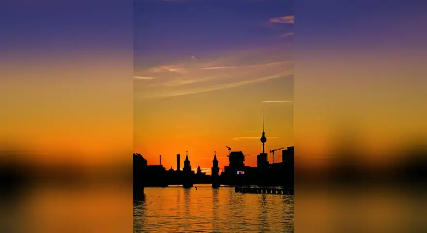 Silhouetten der Berliner Skyline spiegeln sich im Wasser. Ein dramatischer Sonnenuntergang taucht den Himmel in Orange- und Violetttöne.