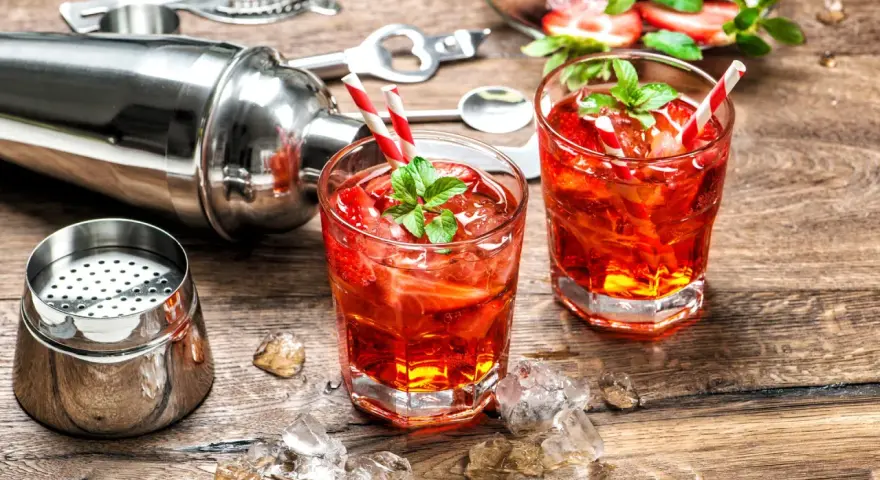 Zwei erfrischende Erdbeer-Cocktails mit Minze und Eiswürfeln, bereit zum Genießen. Ein Shaker und Bar-Werkzeuge deuten auf das kunstvolle Alkohol mischen hin.