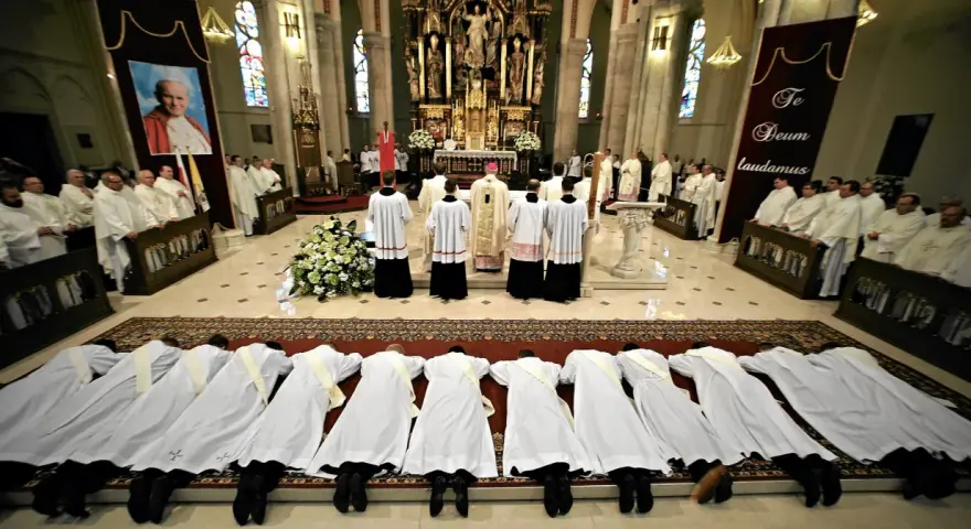 Klerycy w białych szatach leżą na posadzce kościoła, w geście pokory. Obok nich stoją inni duchowni, a w tle widać ołtarz i portret papieża.