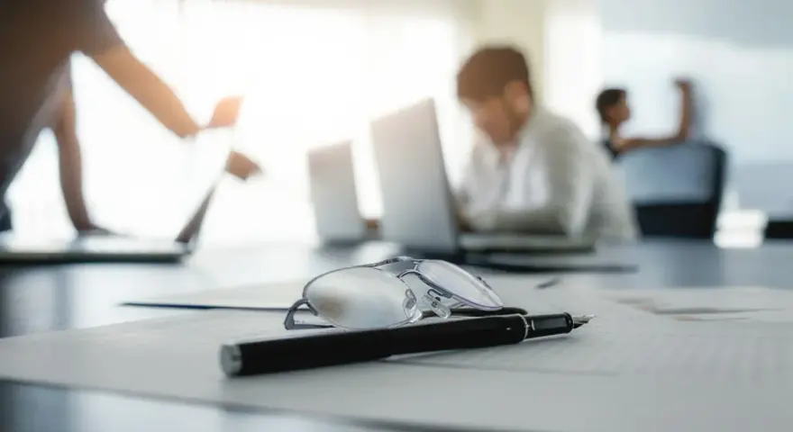 Okulary i długopis na dokumentach, w tle pracownicy analizują nowy kodeks pracy.