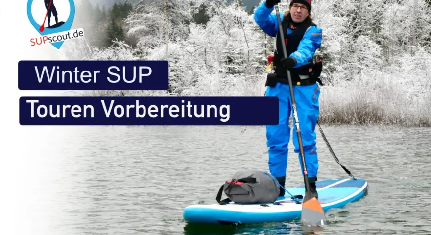 Person im blauen Neoprenanzug bereitet sich auf eine Winter SUP Tour vor. Die Landschaft ist mit Schnee bedeckt.