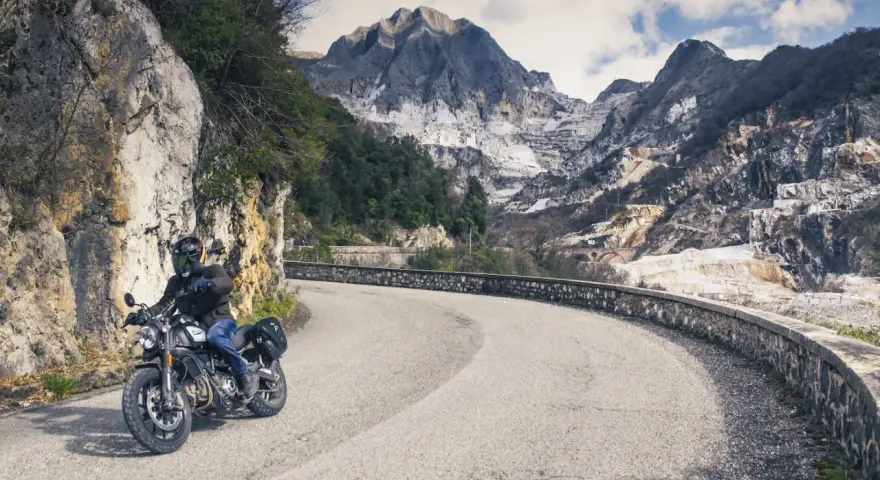 Un motociclista percorre una strada tortuosa tra le montagne della Toscana, con cave di marmo sullo sfondo.