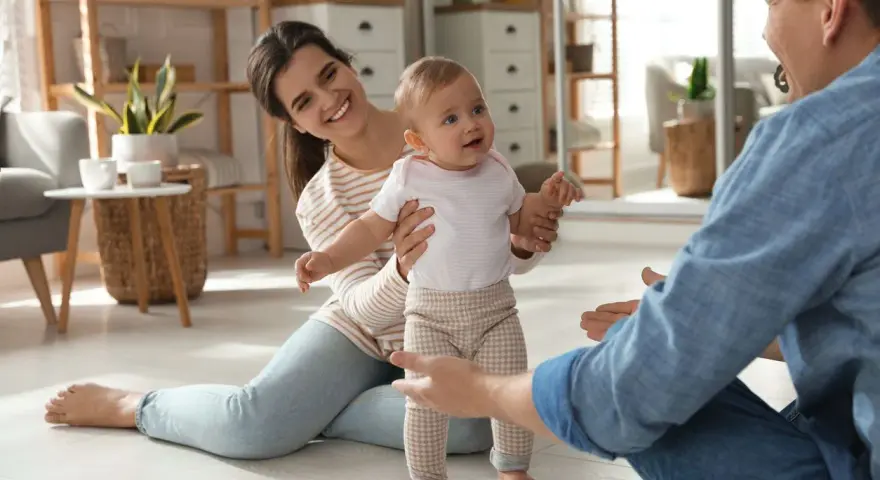 Rodzice zachęcają dziecko do pierwszych kroków. Uśmiechnięta mama i tata z otwartymi ramionami motywują malucha do chodzenia.