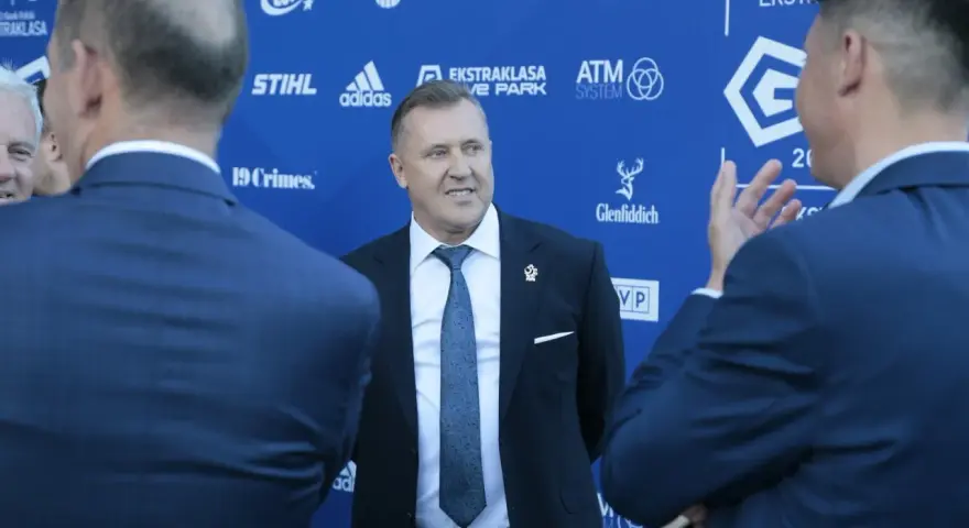 Mężczyzna w garniturze uśmiecha się, stojąc na tle niebieskiej ściany z logotypami sponsorów, być może rozmawiając o tym, ile dostają kluby ekstraklasy.