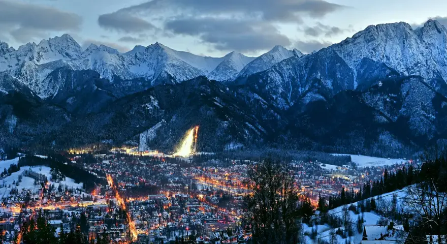 Zimowy wieczór w Zakopanem. Oświetlone miasto u stóp ośnieżonych Tatr, gdzieś w oddali widać stok narciarski. Marzą się bilety na Giewont.