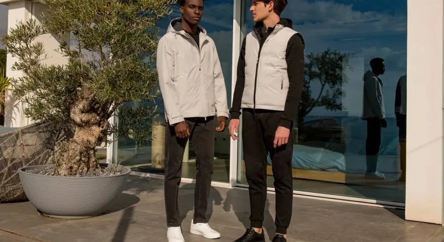 Due uomini con un moderno stile outfit uomo, uno con giacca grigia e jeans, l'altro con gilet grigio e pantaloni sportivi neri.