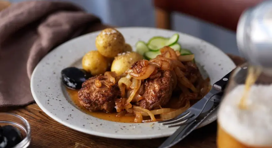 En tallrik med pannbiff, stekt lök, kokt potatis, gurka och en sked med oliver. Ett glas öl hälls upp i bakgrunden. Ett klassiskt pannbiff recept.