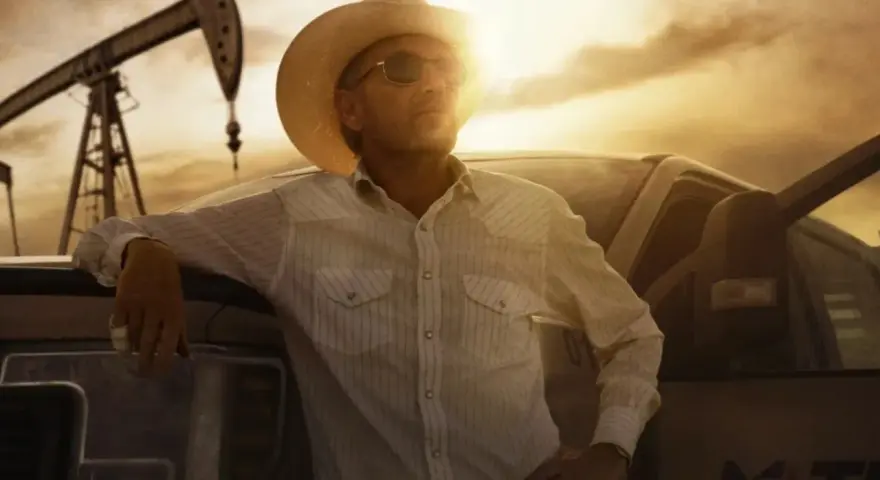 Uomo con cappello da cowboy e camicia a righe appoggiato a un pick-up, con pozzi petroliferi sullo sfondo. Il western significato è qui, tra lavoro e orizzonti infiniti.