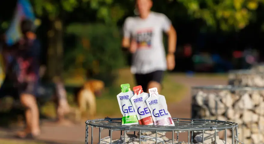 Trzy opakowania żeli energetycznych dla biegaczy: zielone jabłko, cola i truskawka z bananem, na tle biegnącej osoby.