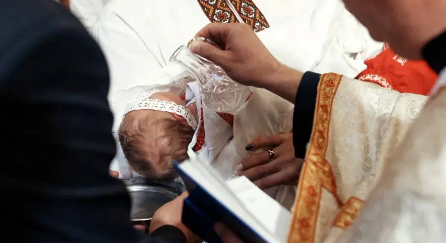 Ceremonia chrztu dziecka. Duchowny polewa główkę niemowlęcia wodą święconą. Warto wiedzieć, ile kosztuje chrzest.