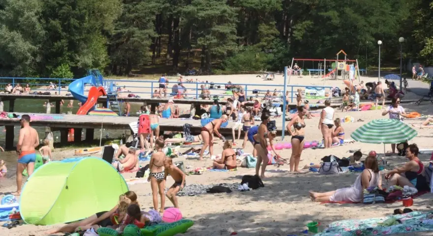 Tłoczna plaża nad jednym z **jezior blisko Wrocławia**. Ludzie opalają się, kąpią i odpoczywają na piasku.