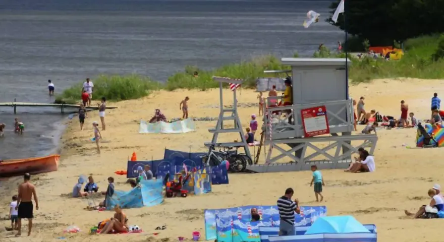 Słoneczna puck plaża tętni życiem. Ludzie opalają się, kąpią i bawią na piasku. W tle widać wieżę ratowniczą i drzewa.