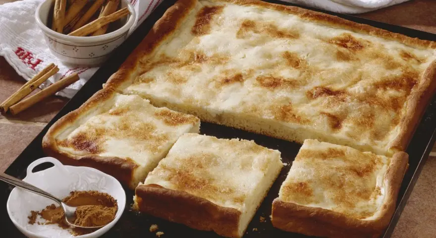 Ein köstlicher Rahmkuchen vom Blech, bestreut mit Zimt und Zucker. Daneben eine Schale mit Zimtstangen und Zimtpulver.