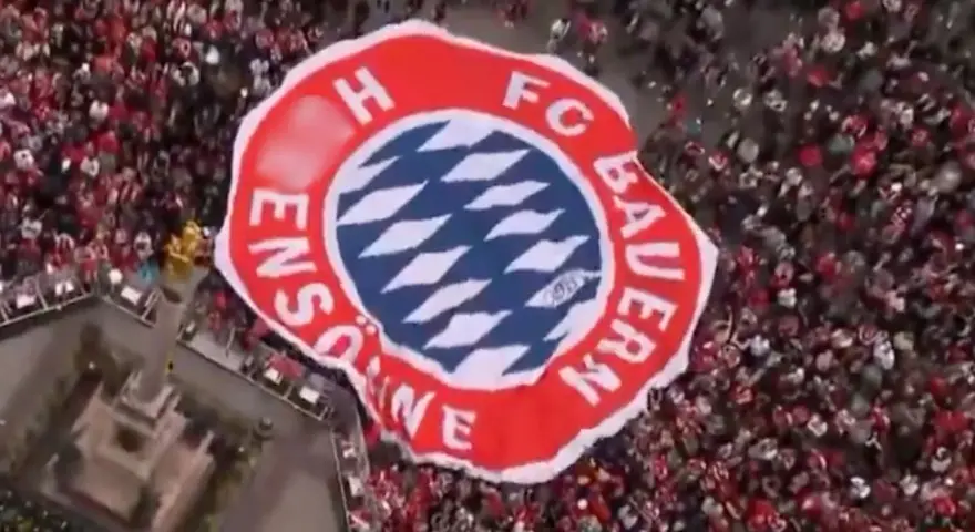 Ogromna flaga z herbem FC Bayern Monachium niesiona przez tłum kibiców.