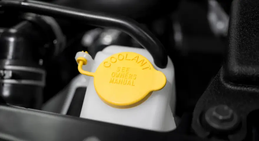 Close-up of a car's coolant reservoir with a yellow cap. The cap reads "COOLANT SEE OWNERS MANUAL," indicating where to find instructions on how to put coolant in car.