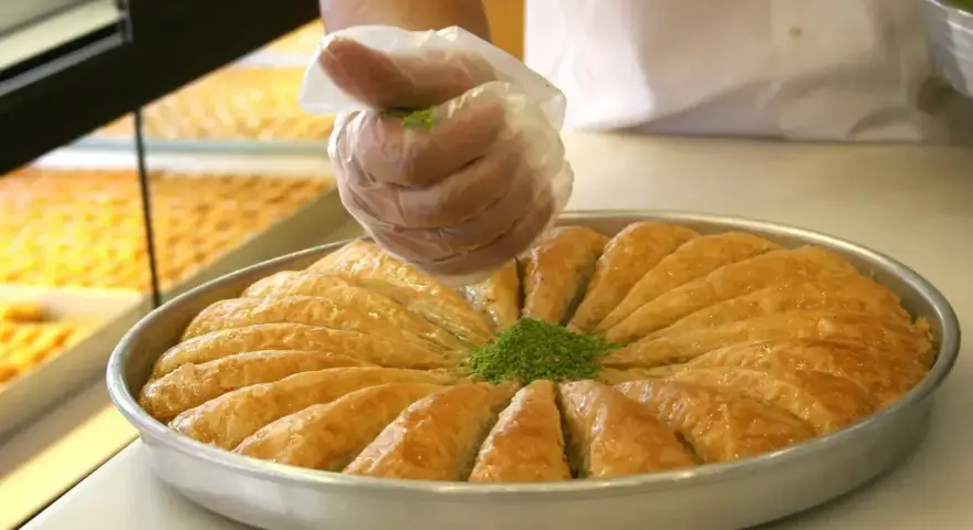 Ręka w rękawiczce posypuje zielonymi pistacjami okrągłą blachę z baklavą. To właśnie baklava co to za deser!