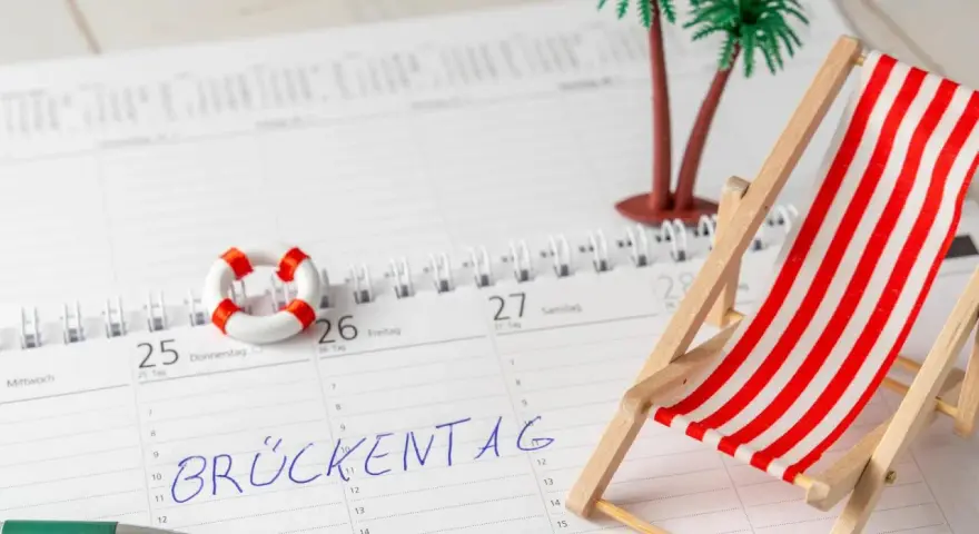 Kalenderblatt mit "Brückentag" markiert. Mini-Strandstuhl, Palmen und Rettungsring deuten auf Pfingstferien 2026 in Bayern hin.