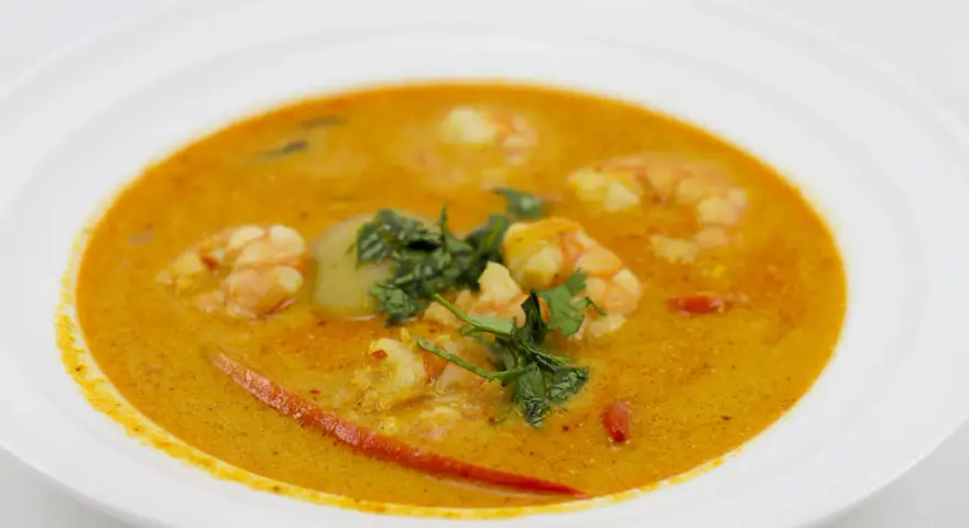 Aksamitna zupa tom yum z krewetkami, świeżą kolendrą i kawałkami chili na białym talerzu.