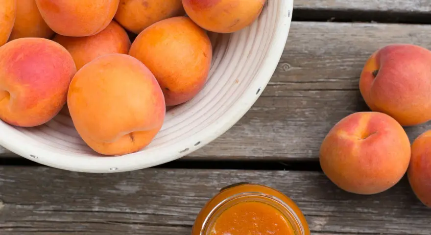 Ein Korb voller reifer Marillen, daneben ein Glas Marillenmarmelade. Was sind Marillen? Süße Früchte, perfekt für Marmelade!