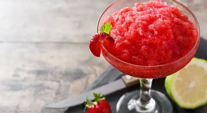 Orzeźwiający strawberry daiquiri, udekorowany plasterkiem truskawki i listkiem mięty, obok połówki limonki i świeżej truskawki.