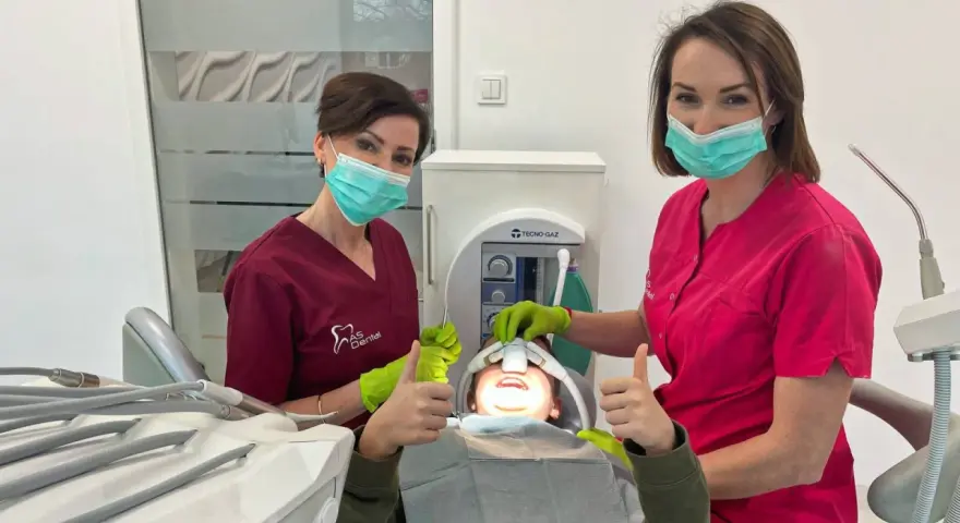 Dwie dentystki w maseczkach i rękawiczkach z uśmiechniętym pacjentem w gabinecie stomatologicznym.
