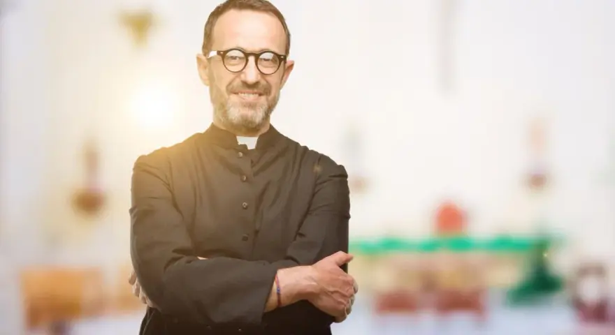 Ein lächelnder evangelischer Pfarrer mit Brille und überkreuzten Armen steht vor einem unscharfen Kircheninterieur.