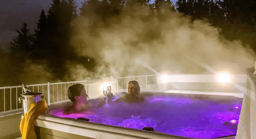 Zwei Frauen stoßen mit Champagner in einem beleuchteten Whirlpool an, umgeben von Dampf und Bäumen. Ein perfektes Wochenende mit Gott.