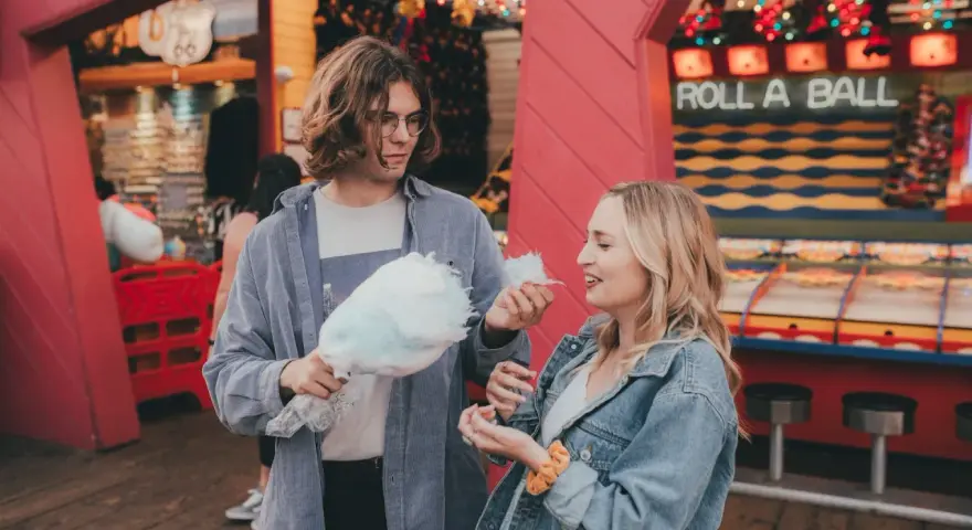Ein Paar teilt sich Zuckerwatte vor einem "Roll a Ball"-Spiel im Freizeitpark. Ihr lässiger Freizeitpark-Outfit passt perfekt zum Tag.