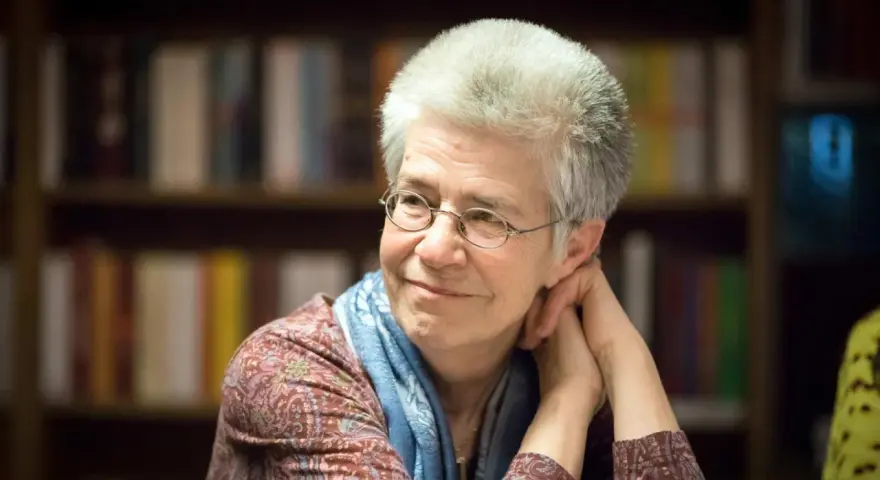 Ältere Frau mit Brille und blauem Schal lächelt nachdenklich vor Bücherregal.