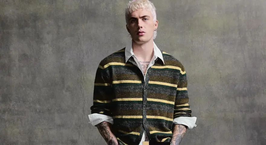 Joven con pelo rubio platino y tatuajes lleva un jersey de rayas verdes y amarillas, mostrando que **que jerseys se llevan este año** son los de estilo retro.