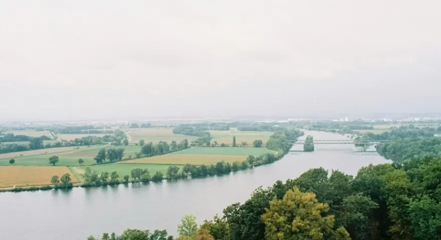 Szeroka rzeka na tle zielonych pól i lasów. W oddali widać most i zabudowania.
