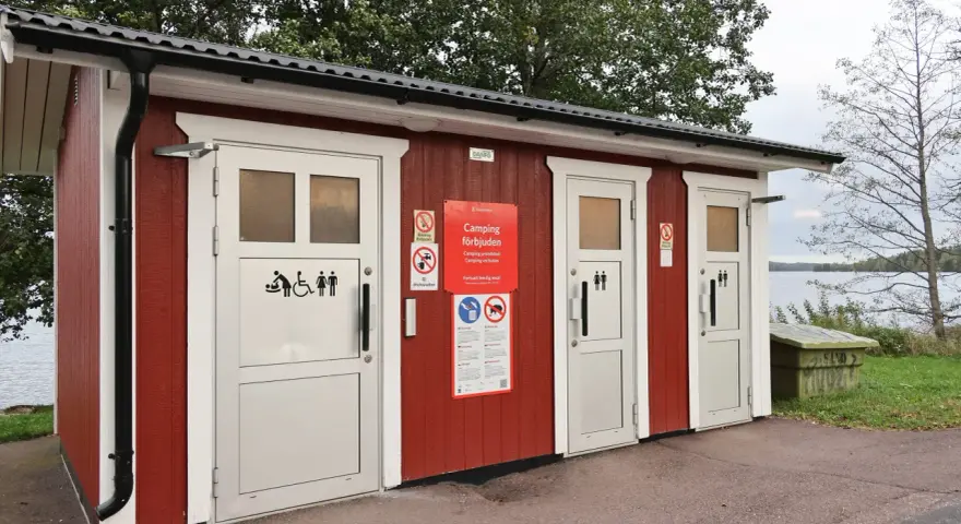 Röd byggnad med toaletter vid en sjö. Skyltar visar att camping är förbjuden. En trevlig rastplats med toalett för resenärer.