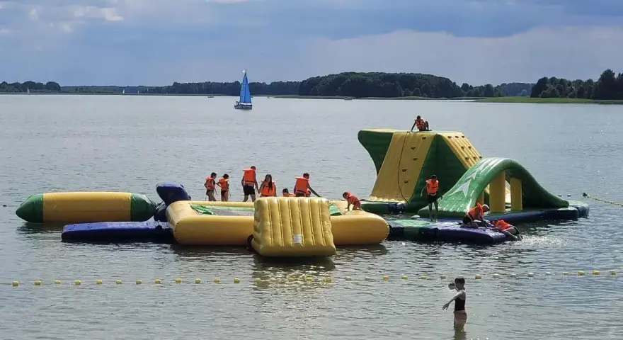 Dzieci w kamizelkach ratunkowych bawią się na dmuchanych wodnych atrakcjach. Idealne miejsce, gdzie na wakacje z dziećmi, z żaglówką na horyzoncie.