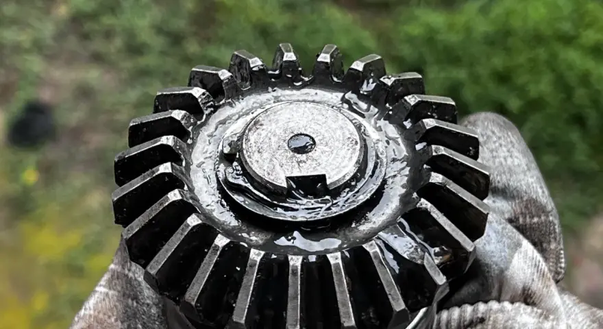 Metalowa przekładnia do mulczera, pokryta olejem, trzymana w rękawicy.