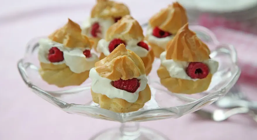 Små petit choux fyllda med grädde och hallon på ett glasfat.