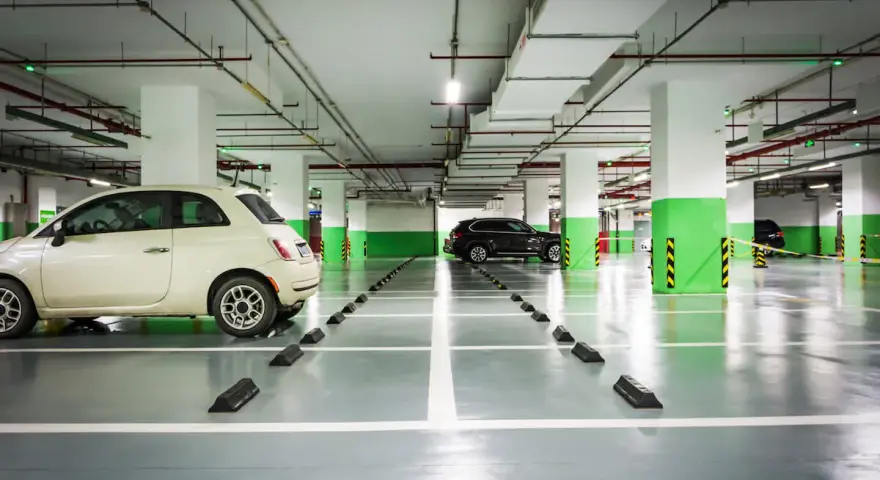 Jasny samochód i ciemny SUV stoją na parkingu podziemnym. Widać wyraźnie wyznaczoną szerokość miejsca parkingowego.